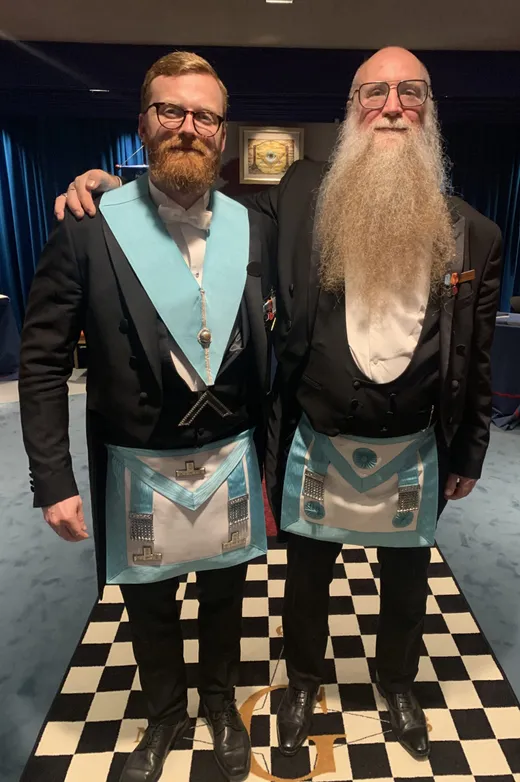 To brødre i kjole og hvidt, den ene med et langt hvidt skæg, står med armen om hinanden